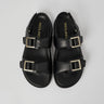 Black Barna leather sandals with gold buckles on a light gray background